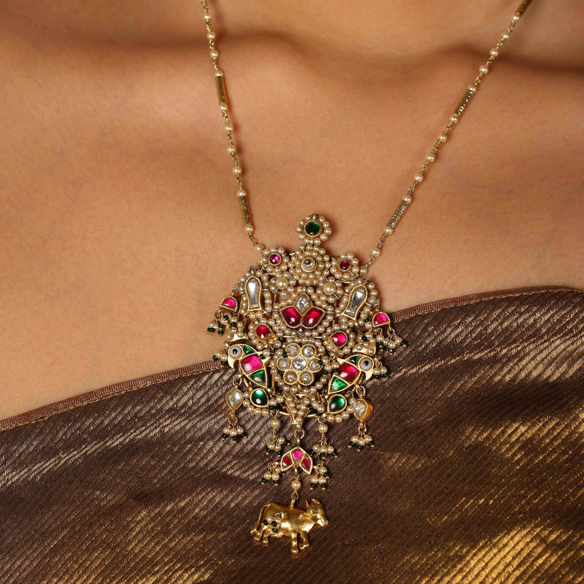 Ornate gold necklace with red and green gemstones on a person wearing a brown garment.